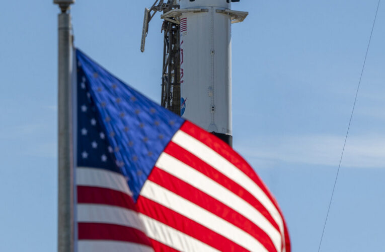 A close-up view of a SpaceX Crew Dragon spacecraft mounted atop a Falcon 9 rocket on the launch pad. The capsule and trunk are connected to ground support hardware, with NASA and mission insignia visible on the rocket’s exterior against a bright blue sky. in the foreground in the American flag.