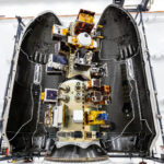 Top‑down view of a SpaceX payload fairing interior showing mounted satellites and hardware arranged along the center structure, surrounded by the curved black fairing walls.