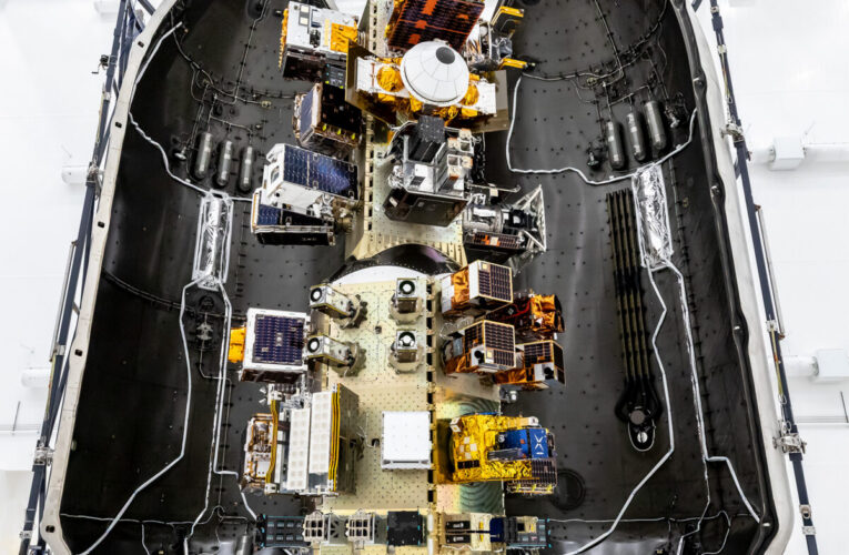 Top‑down view of a SpaceX payload fairing interior showing mounted satellites and hardware arranged along the center structure, surrounded by the curved black fairing walls.